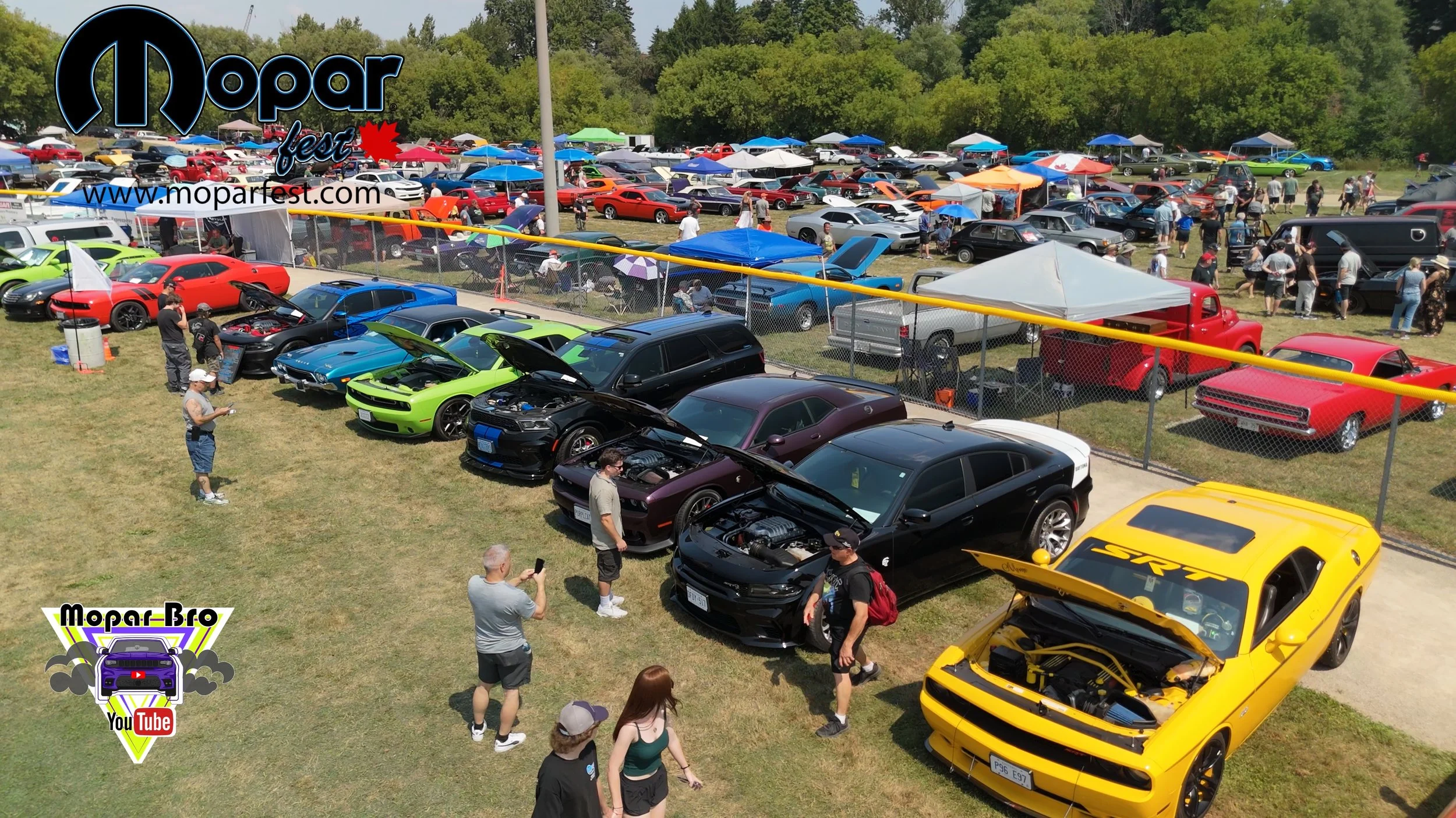 A car show with various cars on display, including a yellow Dodge Challenger with its hood open, some vehicles with their hoods raised, and many spectators walking around and taking photos. There are tents, umbrellas, and a crowd in the background.
