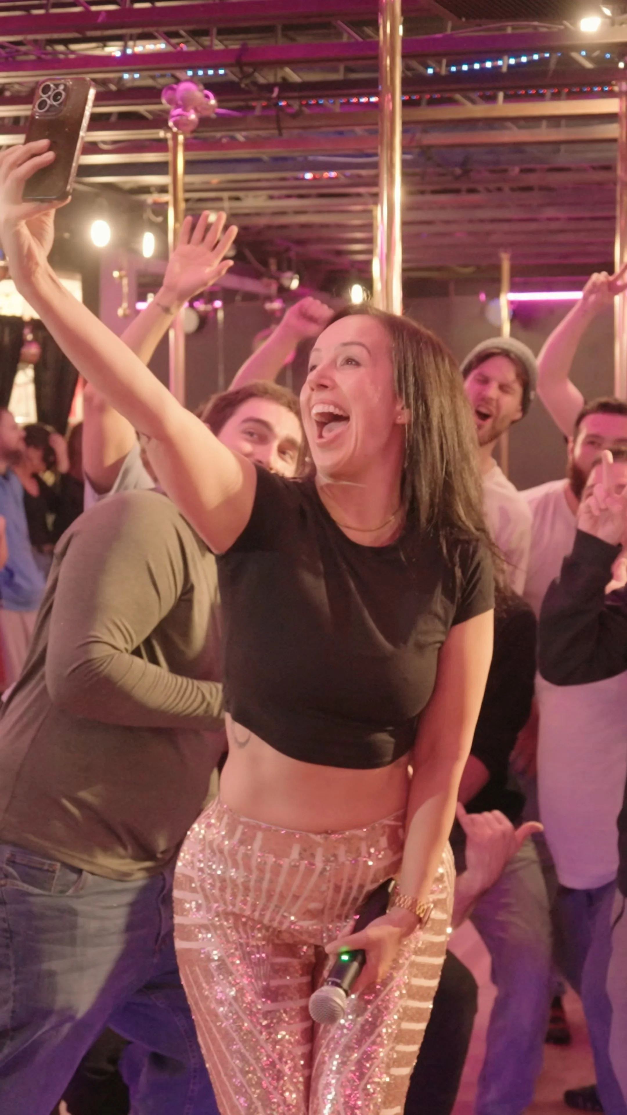 A group of people at a lively party, smiling and having fun, with a woman holding a microphone in her hand and taking a selfie with the crowd.