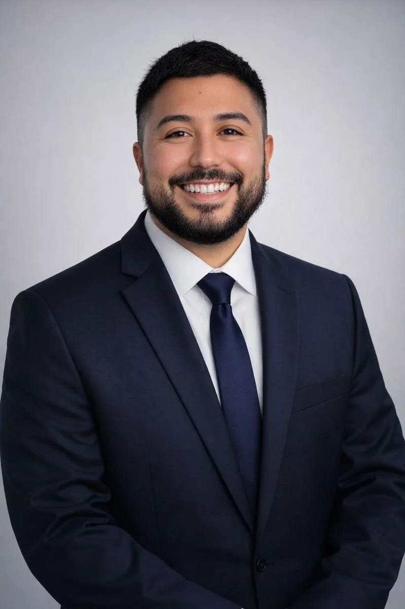 A professional headshot of a man in a dark suit, white shirt, and dark tie, smiling with short dark hair and a beard, against a plain gray background.