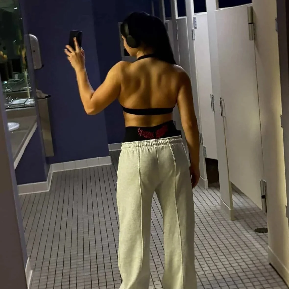 A woman taking a selfie in a bathroom mirror, wearing headphones, a black sports bra, and beige sweatpants.