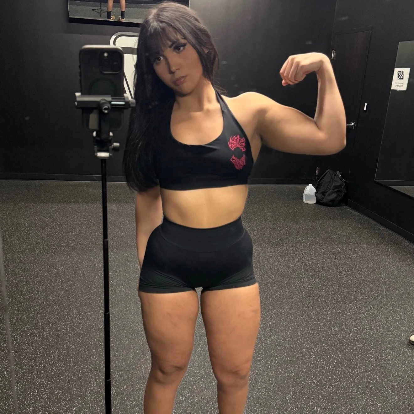 A woman taking a mirror selfie in a gym, flexing her right arm, wearing a black sports bra and black shorts.