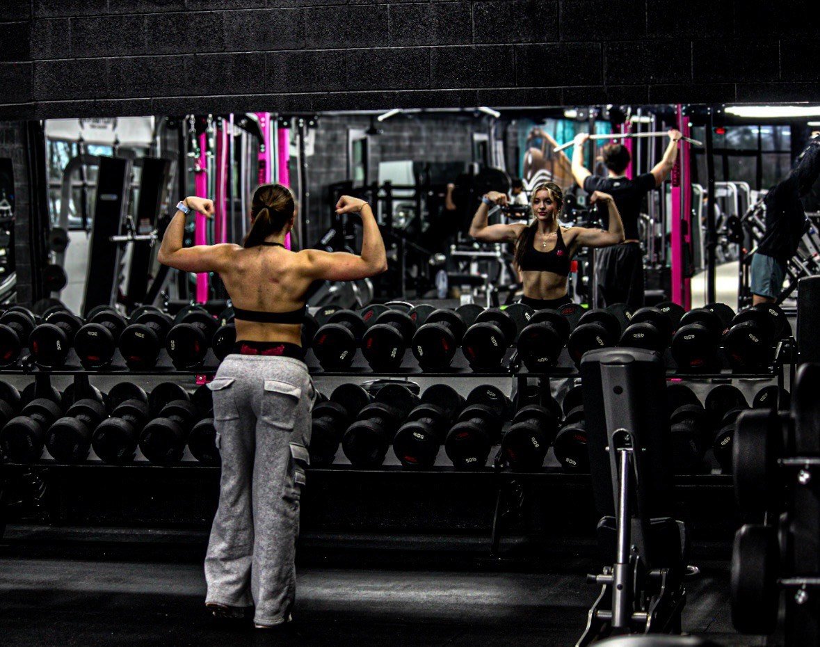 A woman at a gym flexes her biceps in front of a mirror, while her reflection shows her flexing, with dumbbells and gym equipment in the background.