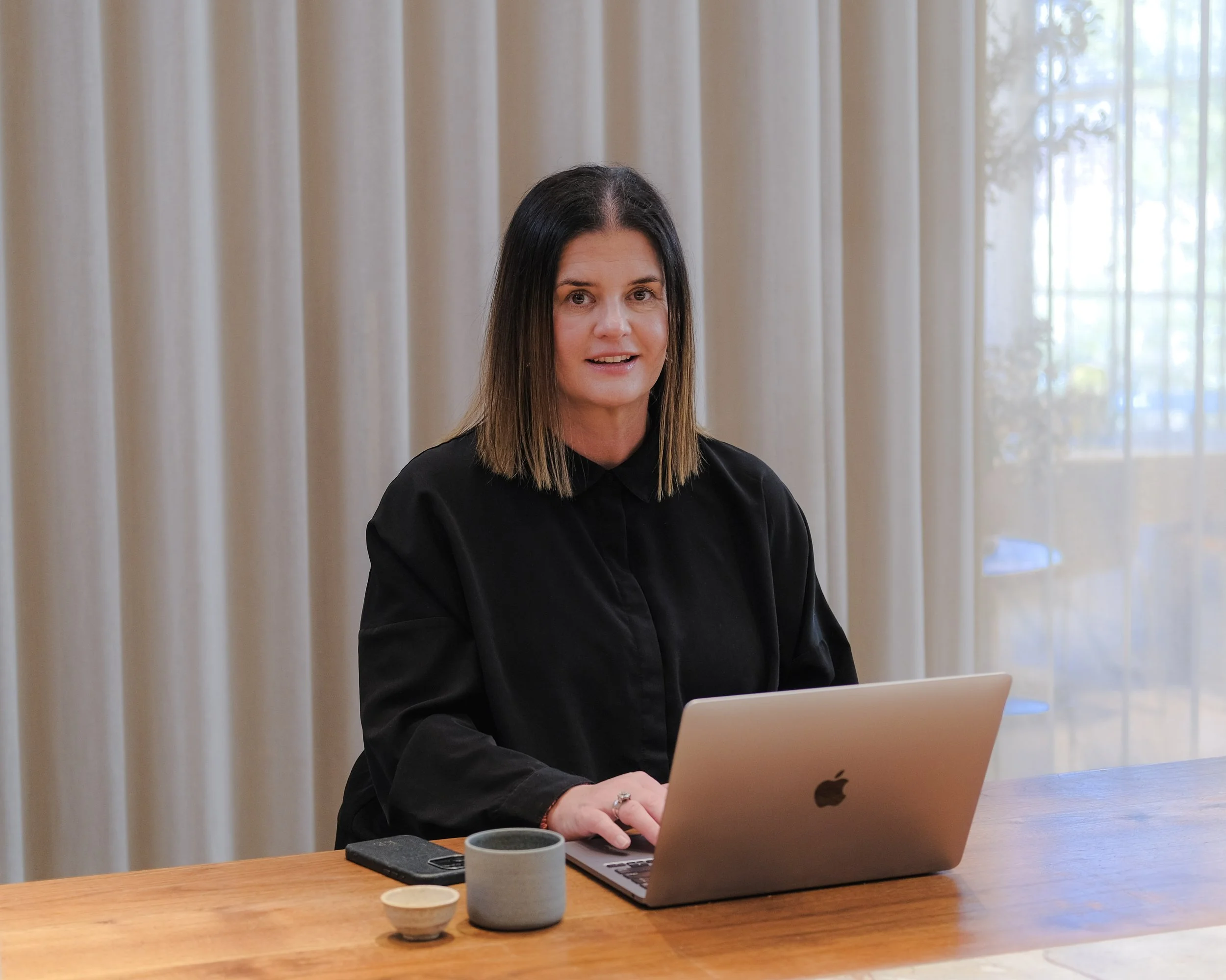 A woman with shoulder-length brown hair working on a silver laptop at a wooden table. She is wearing a black shirt and has a ring on her finger. There are three ceramic cups in front of her, and behind her are beige curtains and a window with trees visible outside.