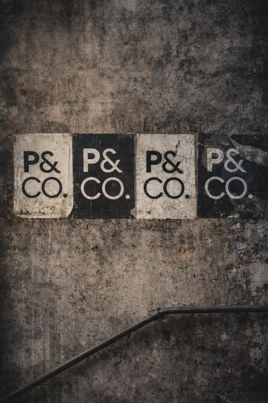 Four worn posters on a rough, dirty wall displaying the text 'P&CO.' in black and white, with a metal railing running diagonally in the foreground.