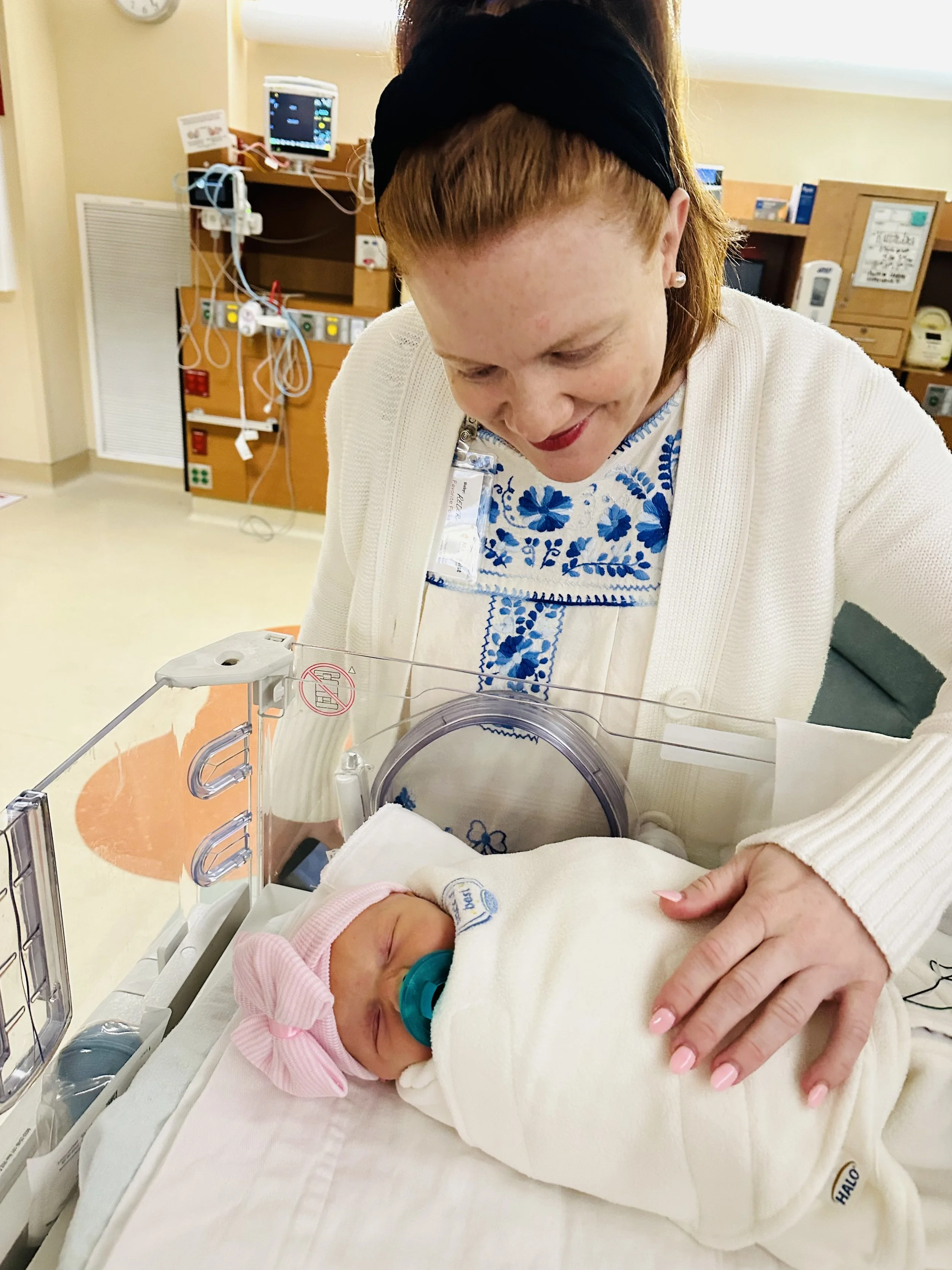 Abigail leaning over her newborn daughter Rosie, swaddled in a hospital bassinet in the NICU