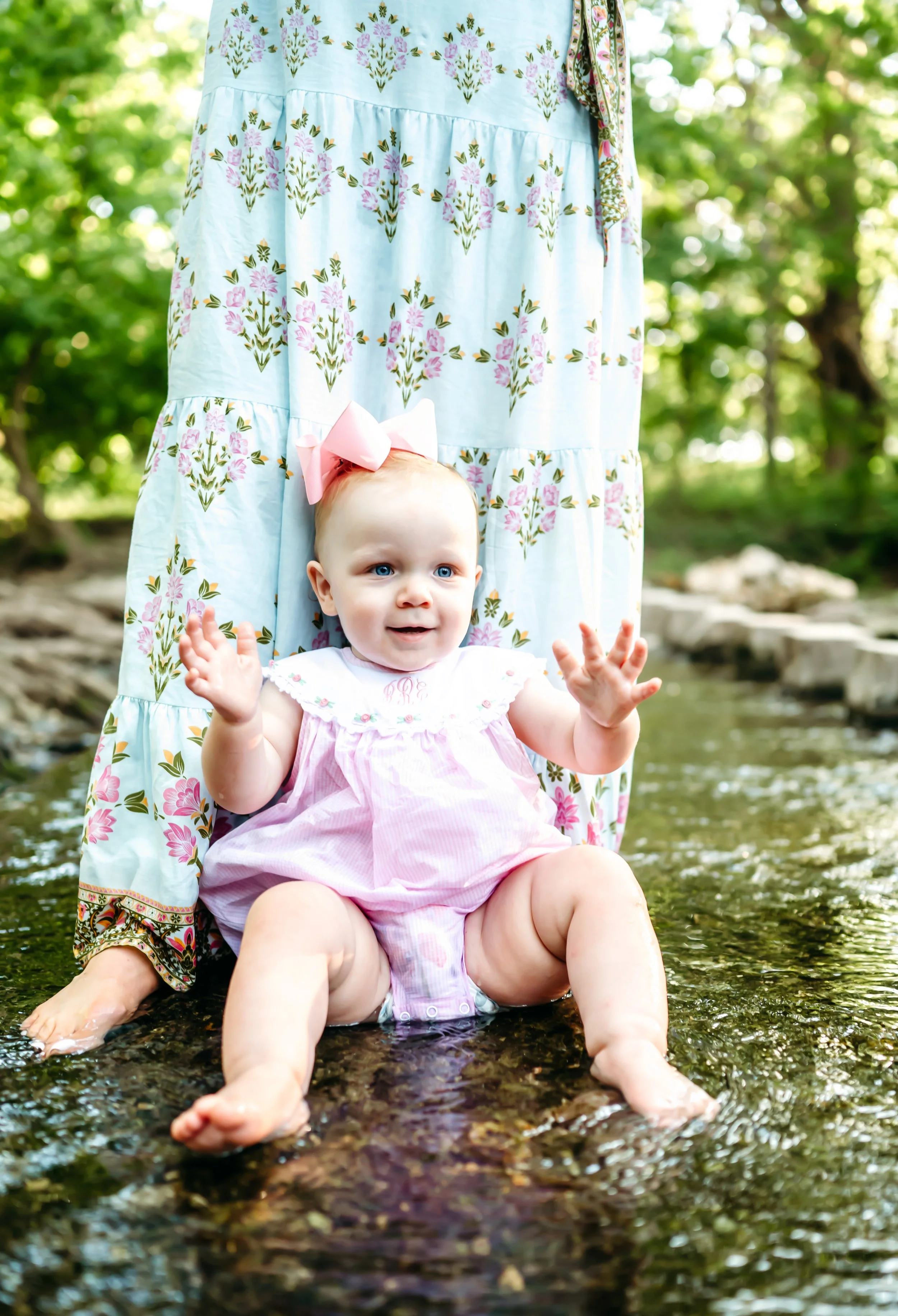 A baby girl in a pink bubble romper and pink bow sits laughing in a shallow Texas creek with her hands raised, her mother standing behind her.
