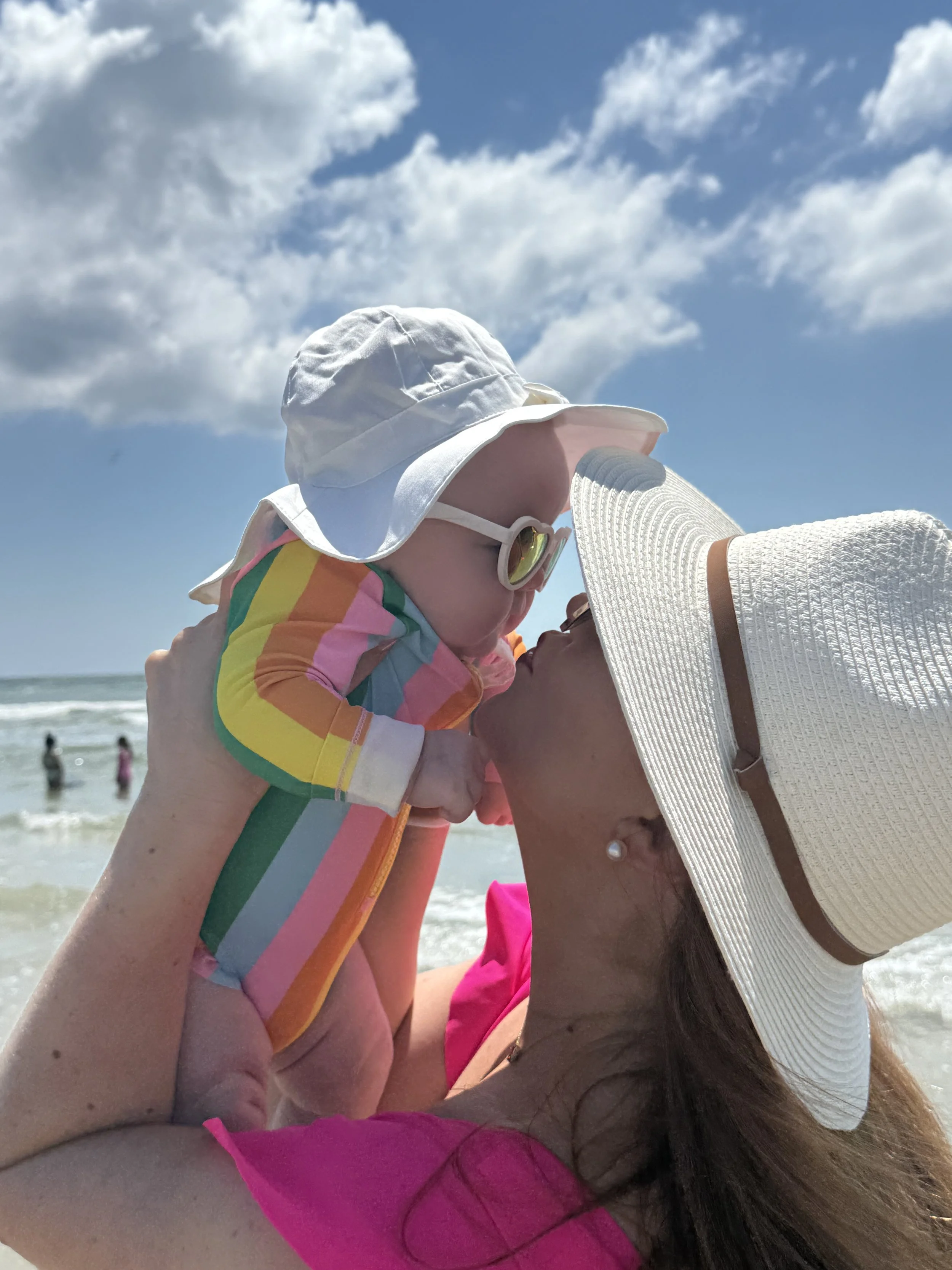 Mom kissing baby on the beach both wearing sun hats for sun protection and sun glasses.