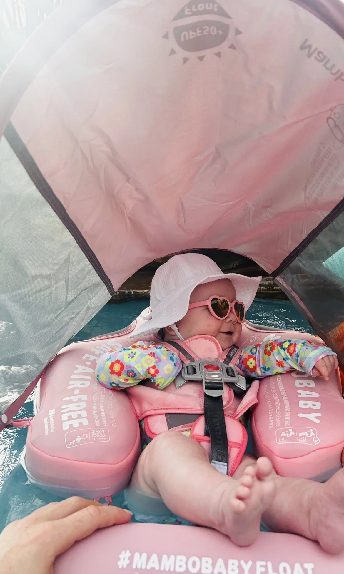 Baby in Mambobaby beach float smiling wearing sunglasses, hat, and rash guard, long sleeve swimsuit in the pool.