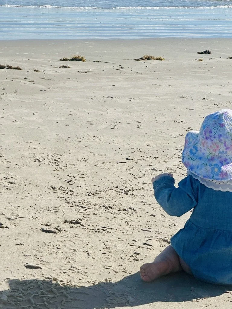 Toddler girl sitting on the beach in Corpus Christi, Texas looking out at the ocean during a peaceful motherhood and coastal family lifestyle moment.