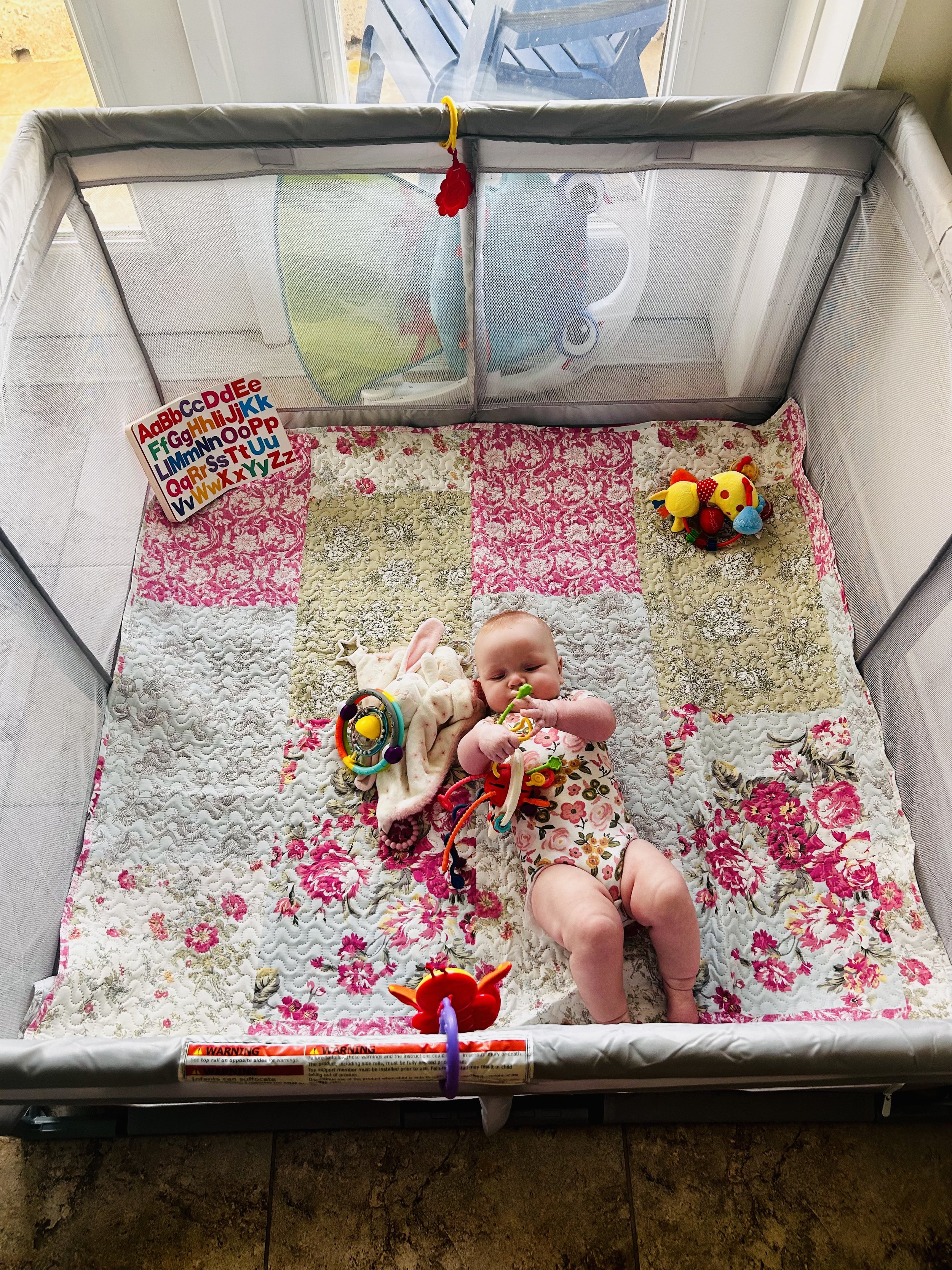 A baby safely laying down in a mesh playpen and playing with a toy.