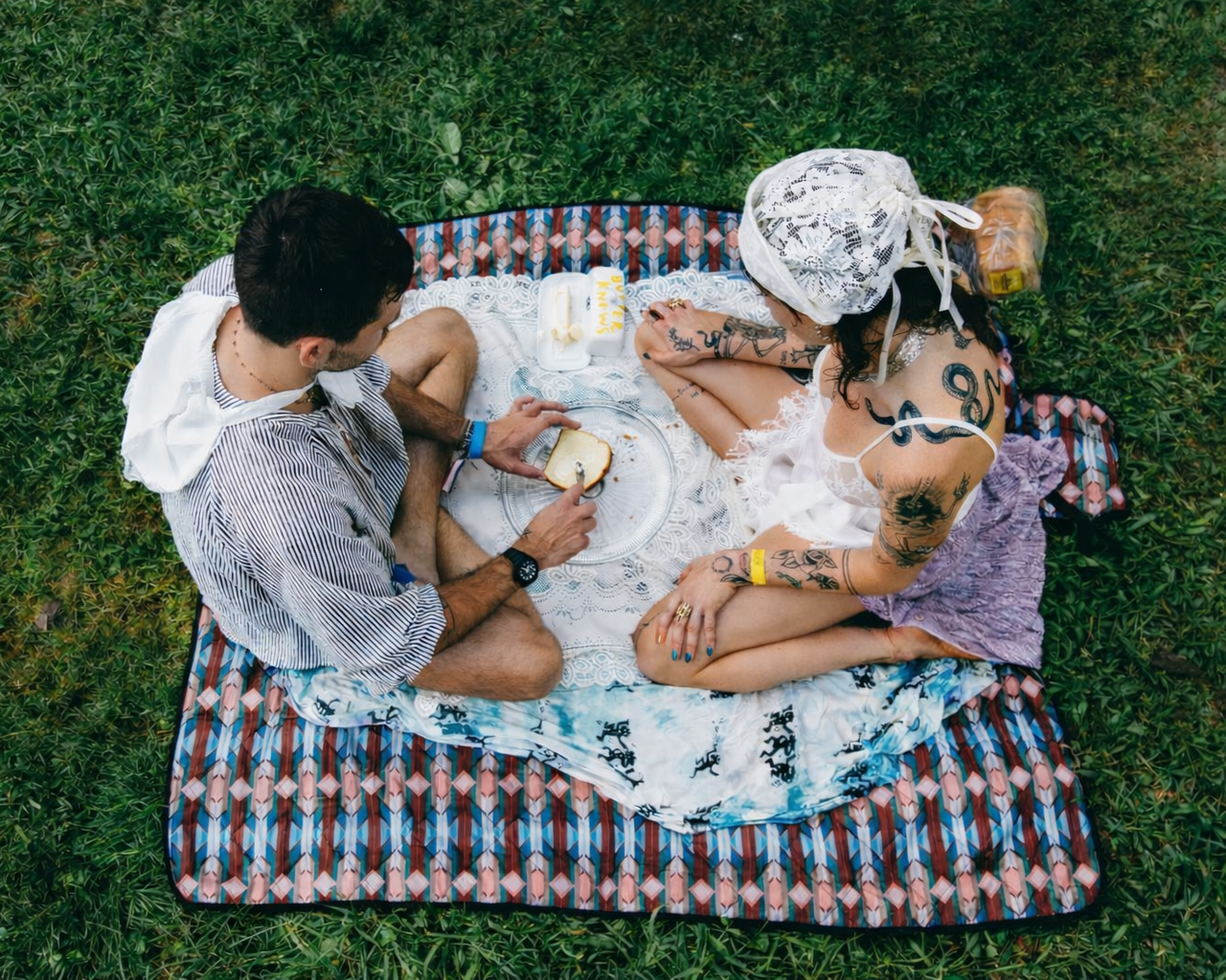 A man and a woman sitting on a blanket in a grassy field. The woman is reading the man's butter oracle. This a a daytime programming event at Churning Man, a butter themed music and art camping festival