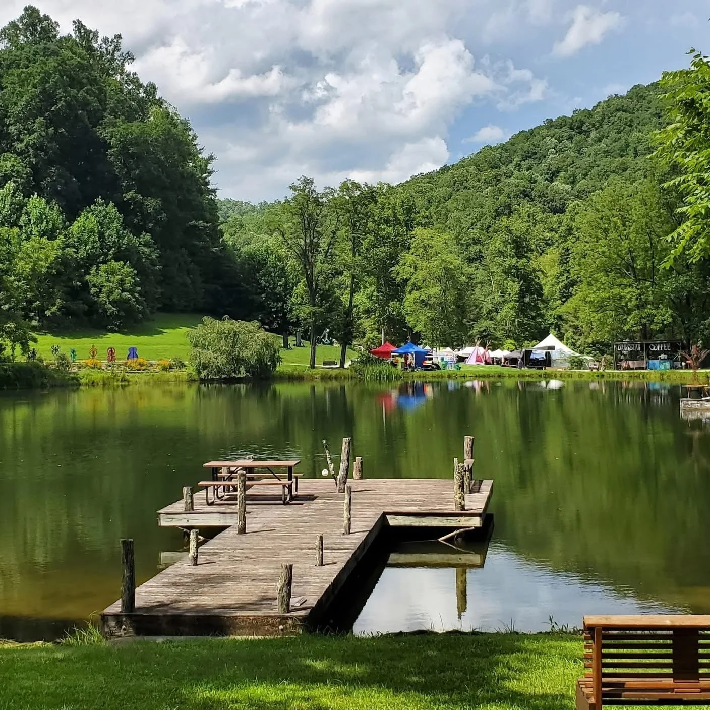 A peaceful lakeside scene at Deerfields, an established outdoor festival venue in Horse Shoe, North Carolina, with a wooden dock extending into the water, surrounded by lush trees and hills. Tents & booths are visible across the lake