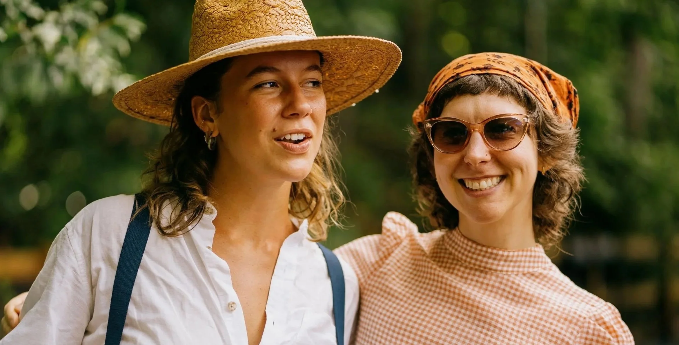 Two women are outdoors, smiling and standing close together, with one wearing a wide-brimmed straw hat and the other wearing sunglasses and a patterned headscarf, in a green, leafy background.