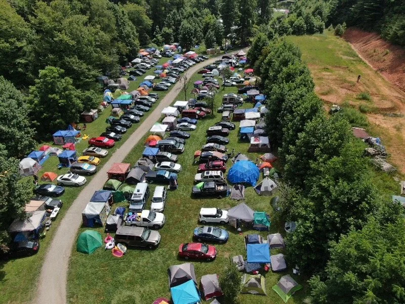 Aerial view of a large outdoor camping area at Deerfields, a outdoor music venue, with numerous tents and parked cars surrounded by trees and greenery.