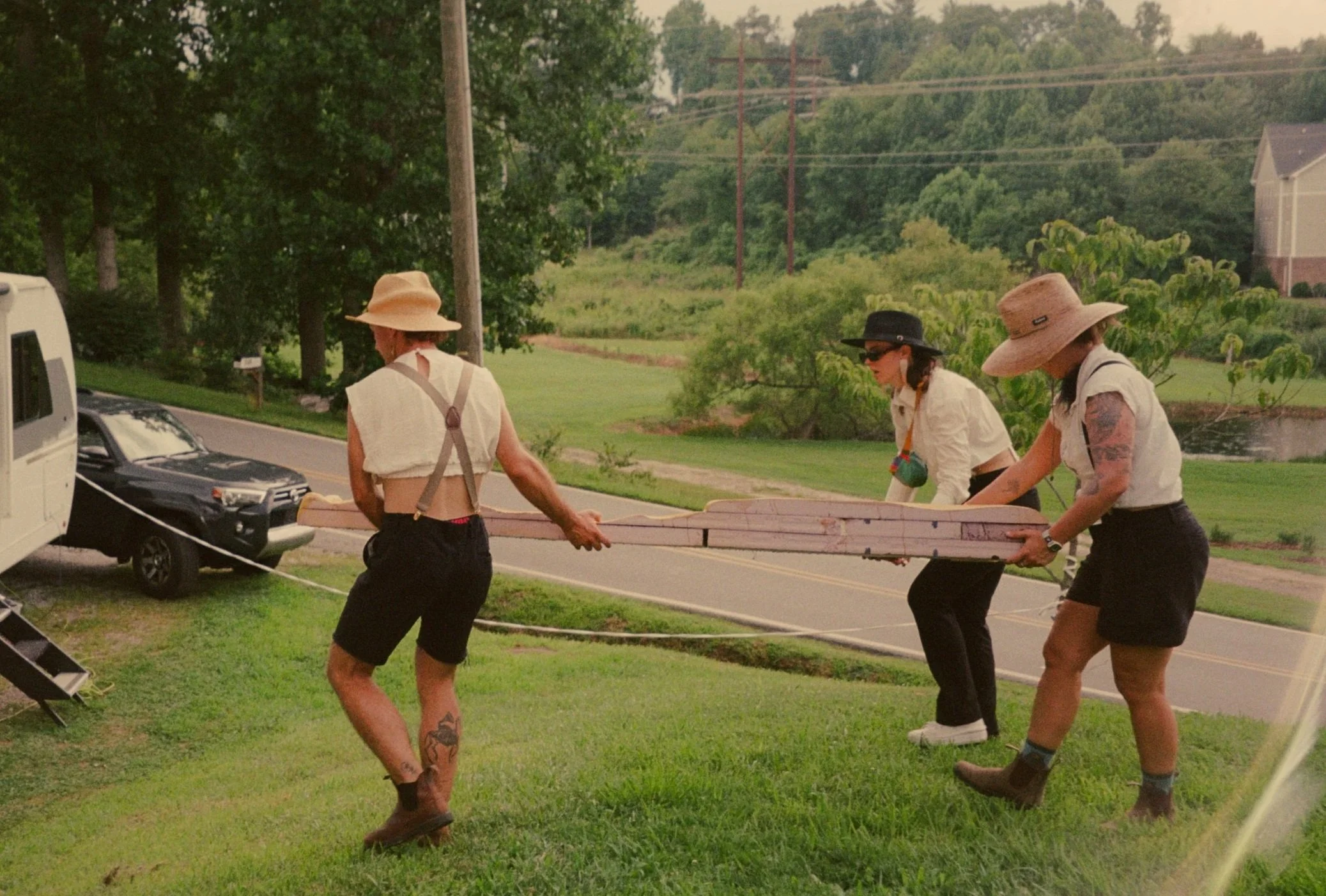 Three women wearing straw hats and old-fashioned clothing are working together as Churning Man volunteers to carry a long wooden plank. They are giving their time and energy to help put on a magical two day camping festival in North Carolina