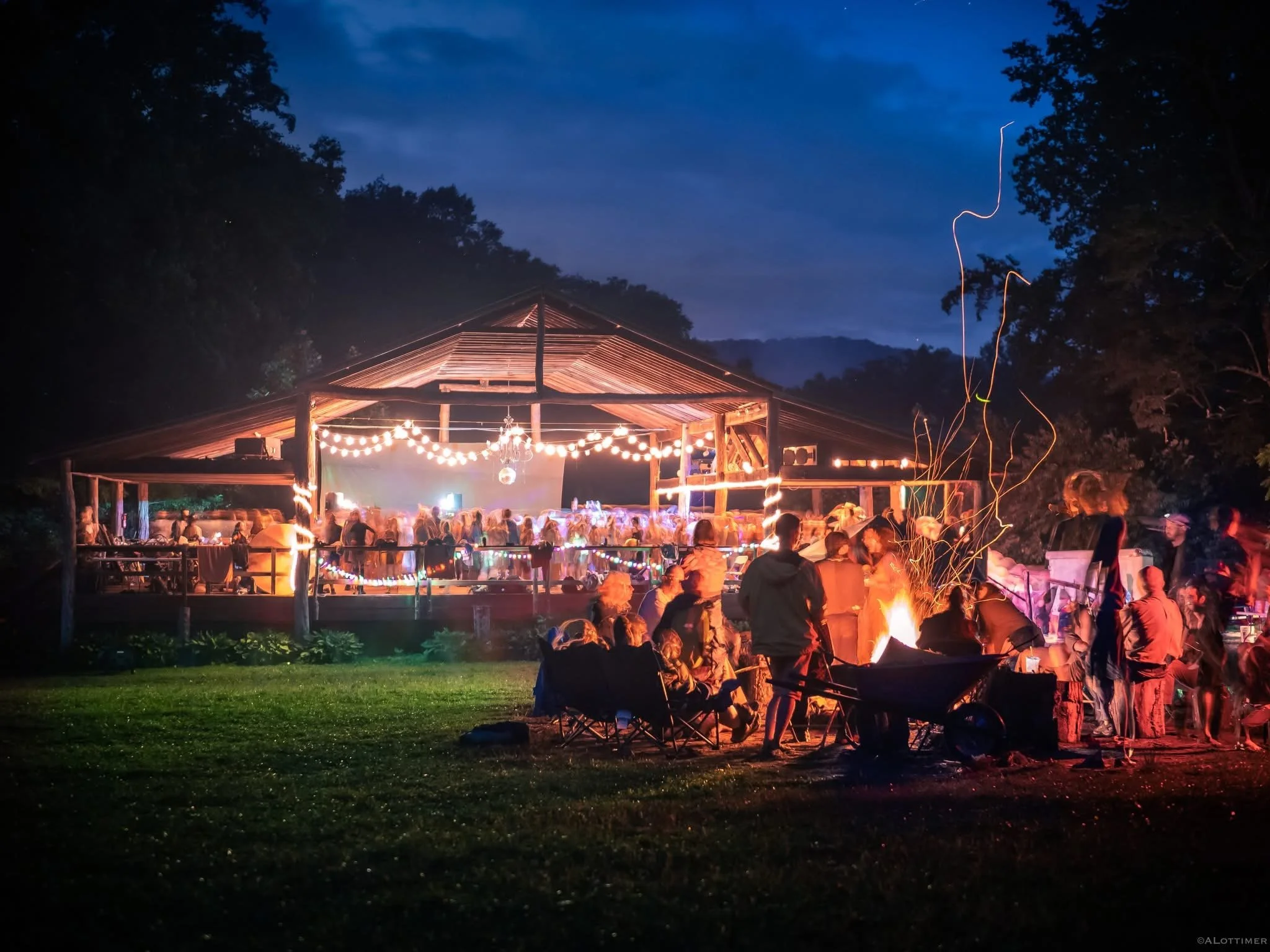 Nighttime outdoor festival scene at at Deerfields in North Carolina. The space is warmly lit, with string lights, a wooden pavilion, and a bonfire with people socializing and dancing.