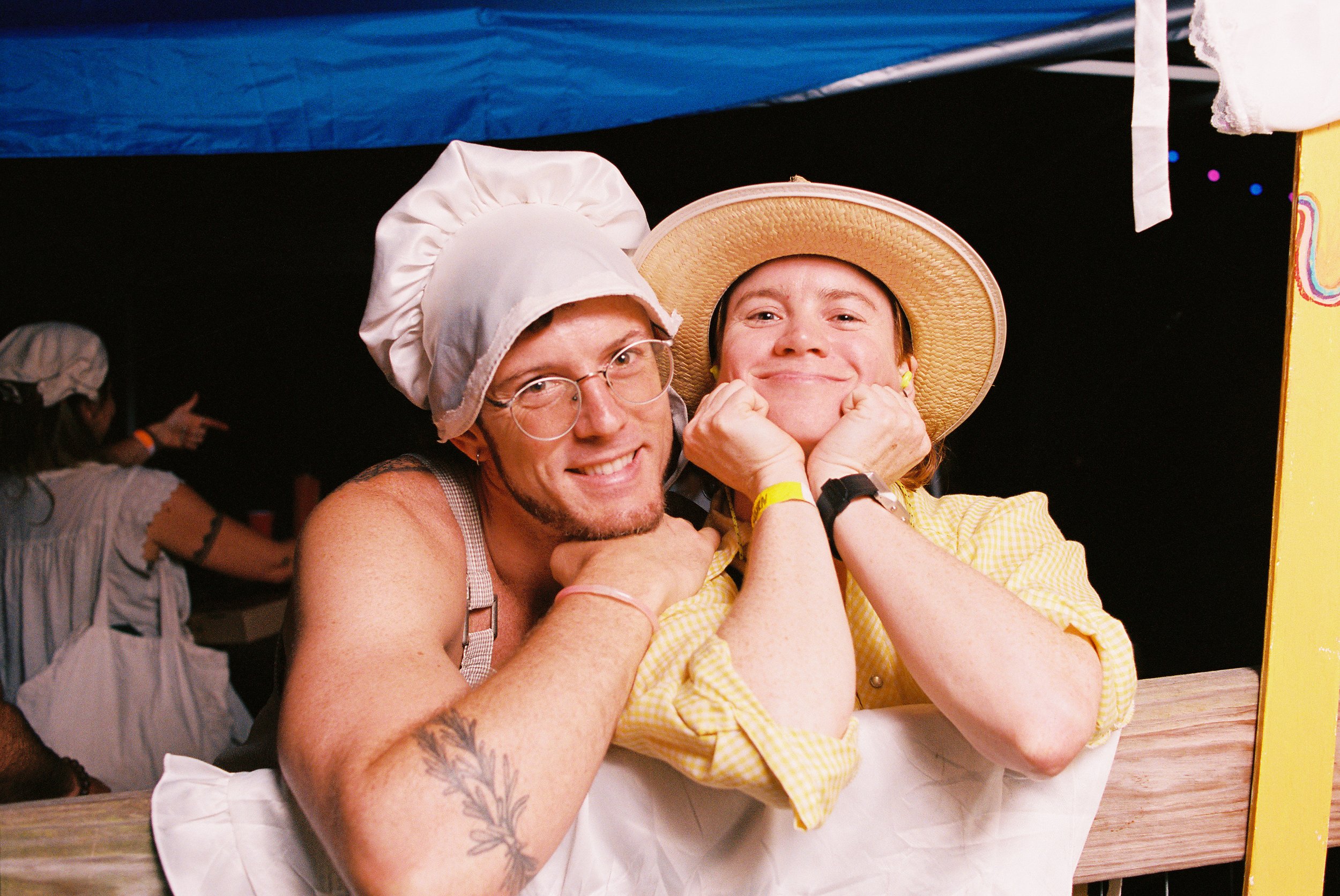 Two people dressed in old-timey buttery churning attire, smiling and posing at an outdoor music and art festival, with lots of people smiling, dancing, and laughing behind them