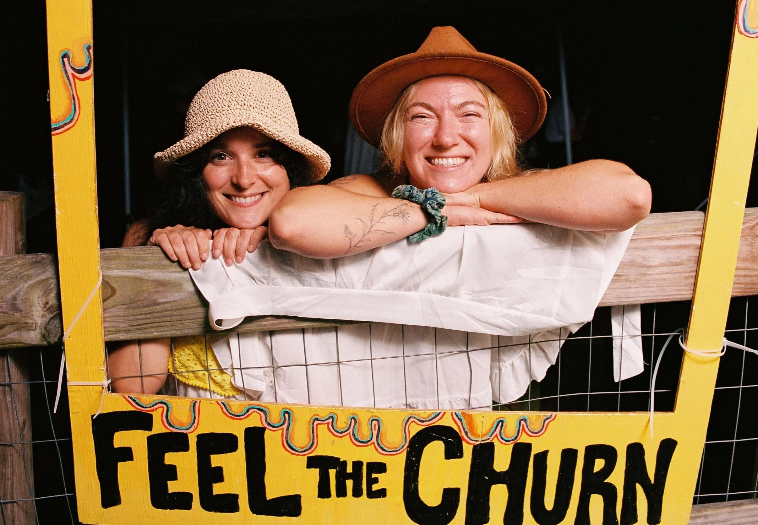 Two women smiling and leaning on a wooden fence at a carnival or fair, with a yellow sign that reads 'Feel the Churn.' They are old-timey attire and are smiling as they make new friends at an art and camping festival