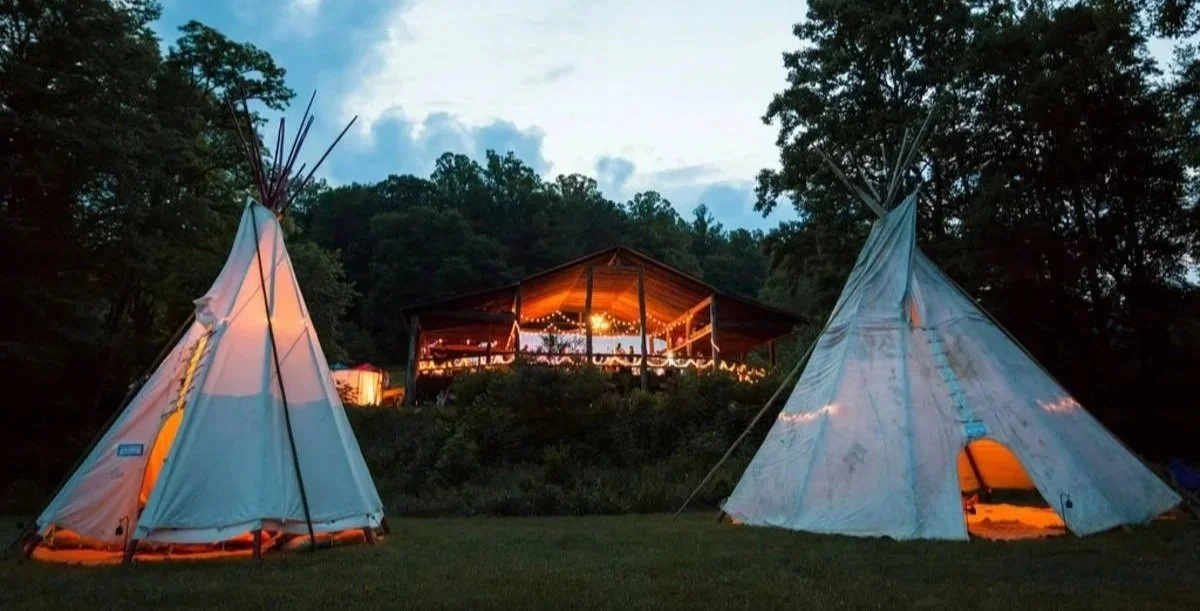 Two teepees with open doors lit from inside, set on grassy area with trees in the background. A wooden building decorated with string lights is visible in the distance against a twilight sky. Deerfields Horse Shoe North Carolina