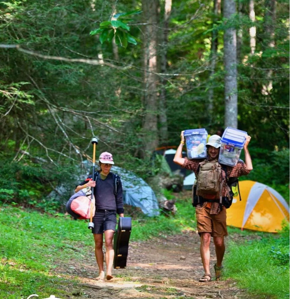 Two people hiking through a forested campsite in North Carolina, carrying camping gear and containers. They are preparing to set up their camp at a 2 day music and art camping festival near Asheville
