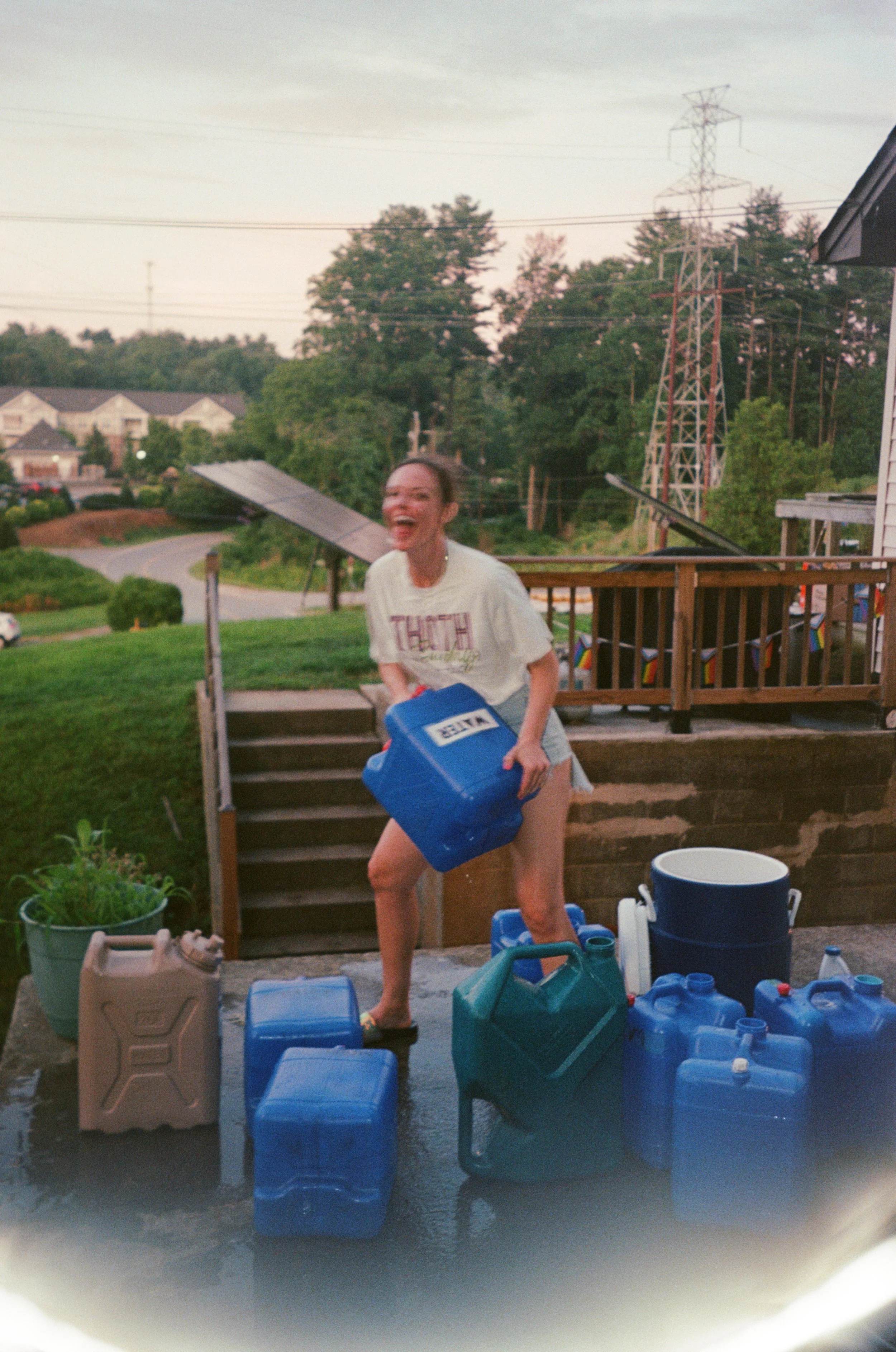 Woman outdoors, holding a blue water container as she volunteers for an outdoor music festival in Asheville North Carolina. She is surrounded by various water containers and jugs, smiling.