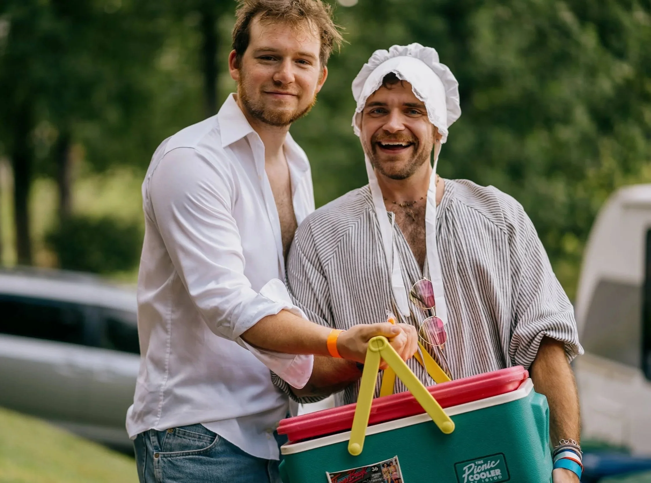 Two men smiling outdoors, one dressed in a striped blouse and a bonnet, holding a colorful cooler, with trees in the background. They are helping one another as they bring beverages to attendees of a music festival in Asheville
