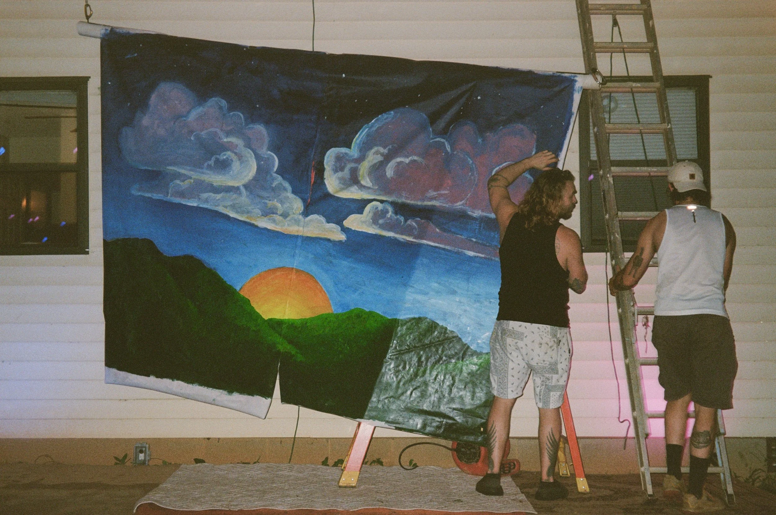 Two men are working on a large landscape painting of a setting sun, mountains, and clouds. One man is standing on a ladder. They are both artists who have brought their creative visions to be showcased at a community powered art and music festival