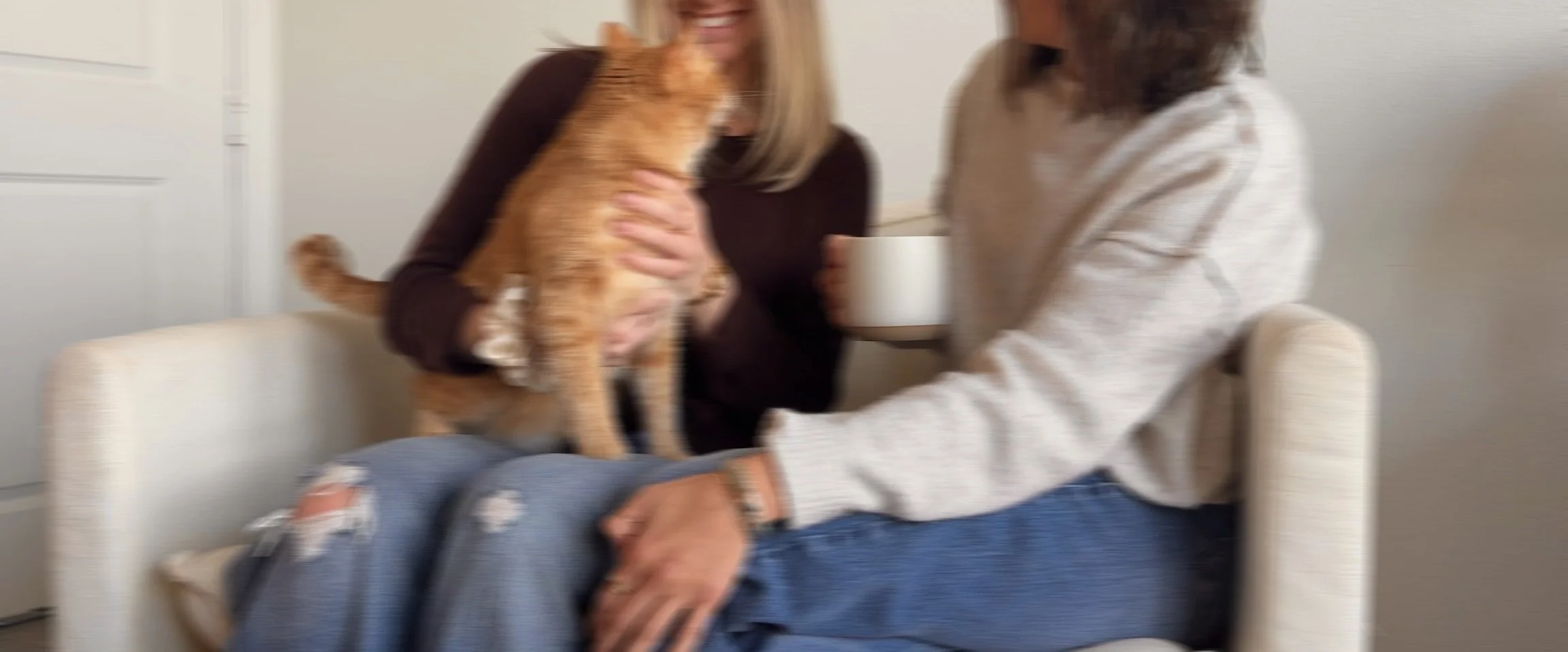 Two women and a cat in a living room. One woman is sitting on a sofa, the other woman is holding the cat and a coffee mug. The cat is an orange tabby. The women are smiling and engaging with each other.