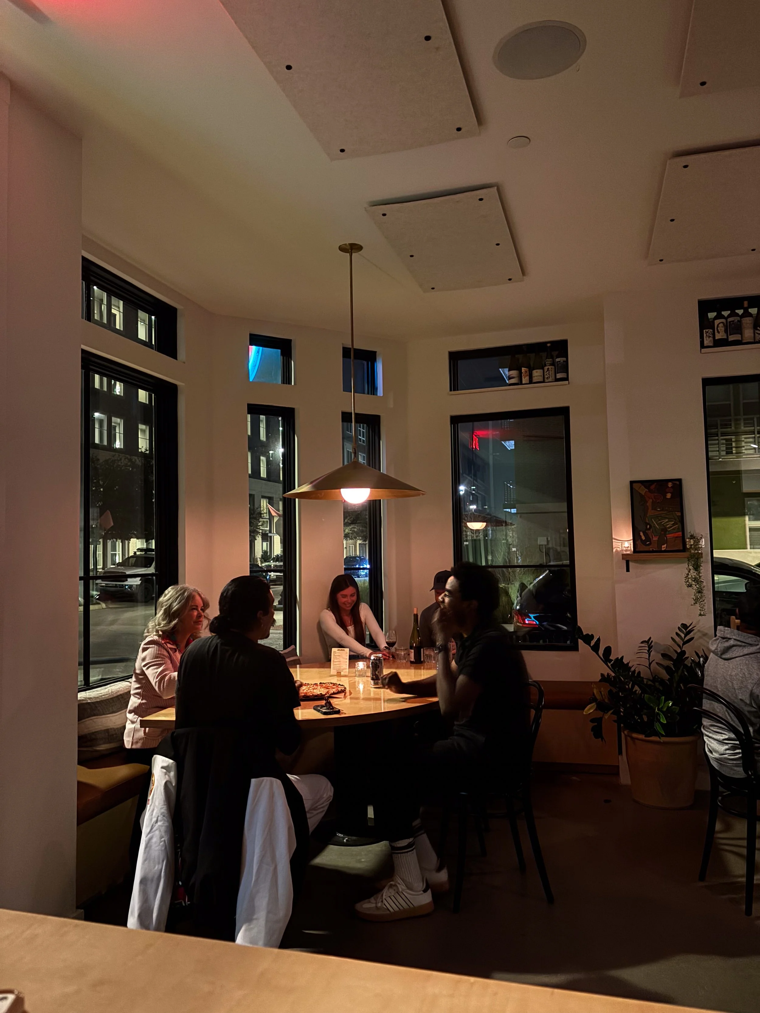 A group of four people sitting around a table in a cozy, modern restaurant at night, enjoying drinks and conversation, with large windows and warm lighting.