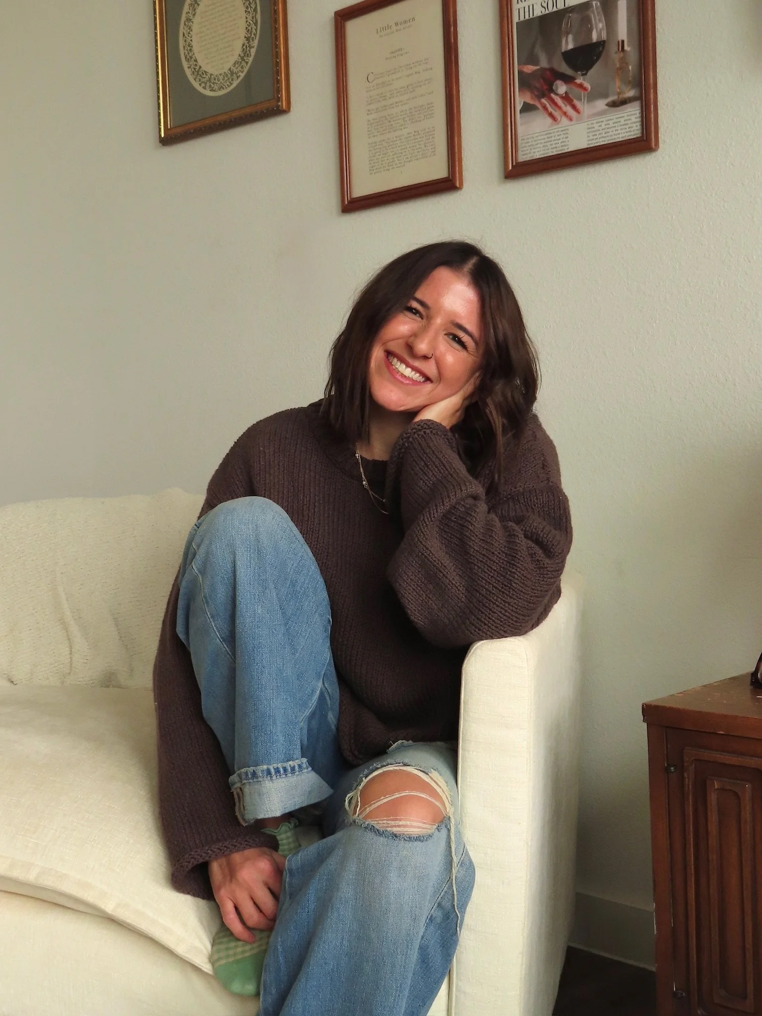 A young woman with shoulder-length dark hair smiling and sitting on a cream-colored couch. She is wearing a brown oversized sweater, ripped jeans, and green socks. She has her left hand resting on her cheek. Behind her are framed magazine pages and articles on a light-colored wall.