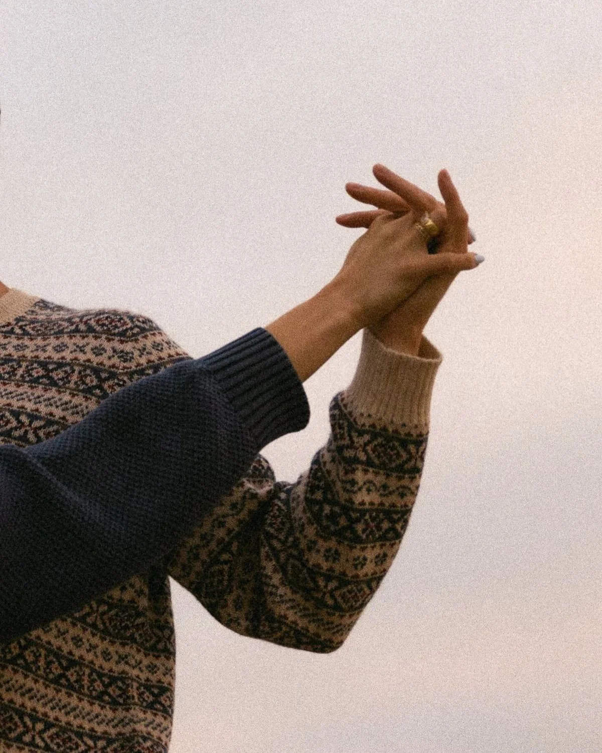 Two hands intertwined, one in a beige and brown patterned sweater and the other in a dark knitted sleeve, against a plain background.