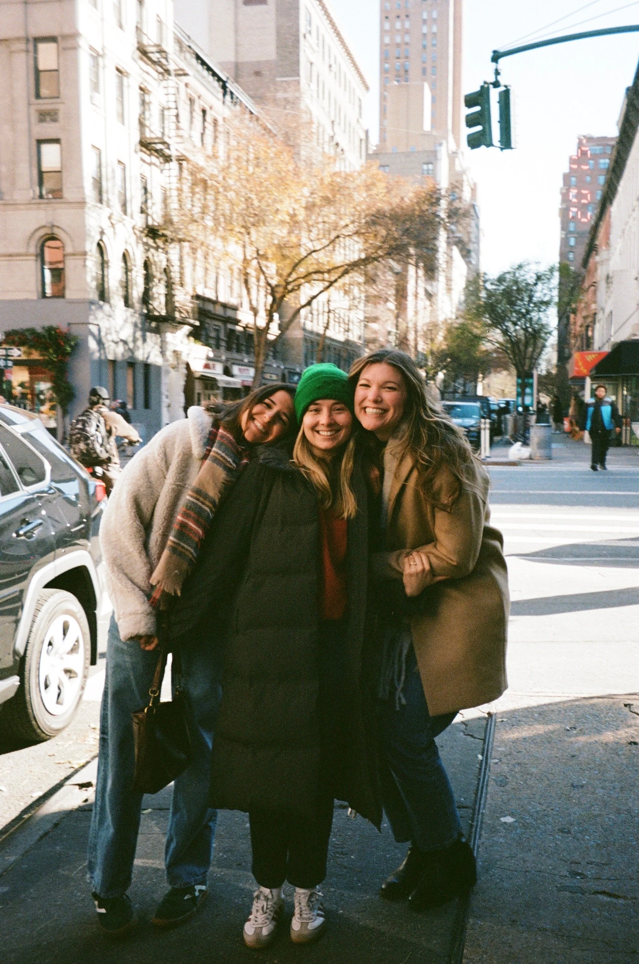 Three women smiling and hugging on a city sidewalk in autumn, with buildings, trees, and cars in the background.