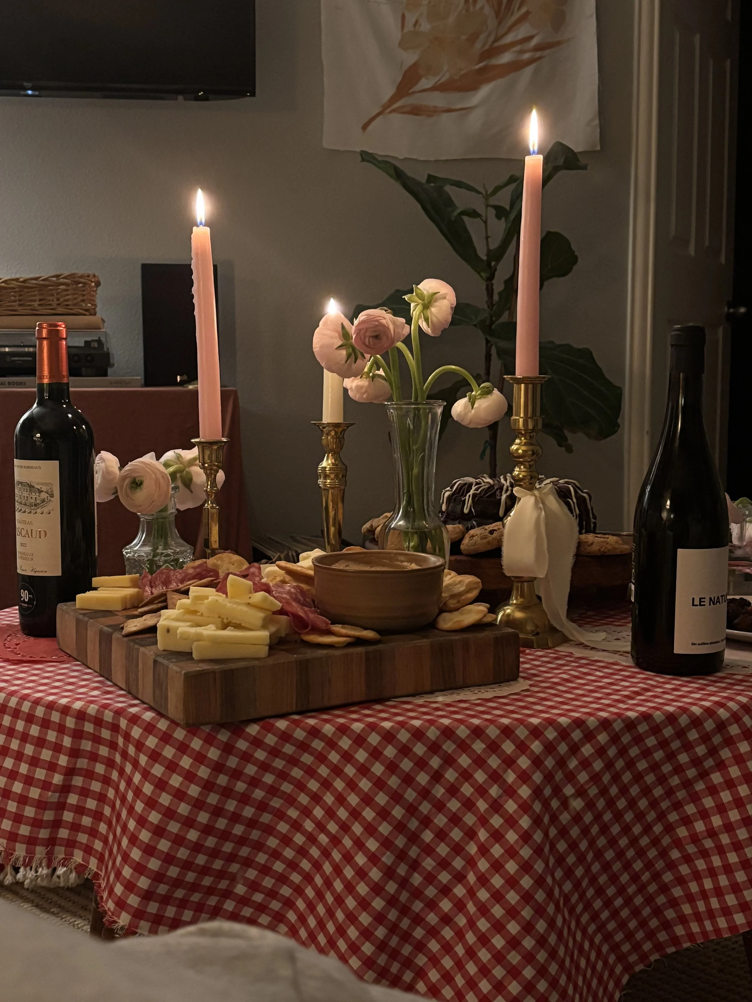 A table with a red and white checkered tablecloth, decorated with pink candles, flowers, wine bottles, and a charcuterie board with cheese, meats, and crackers.