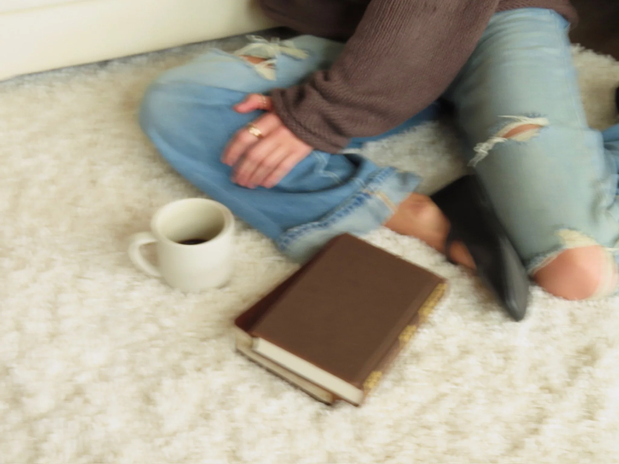 Person sitting on a carpeted floor, wearing ripped jeans and black shoes, with a book, a white mug of coffee or tea, and a black girl. The person is resting their head on their arm, with their face partially visible.