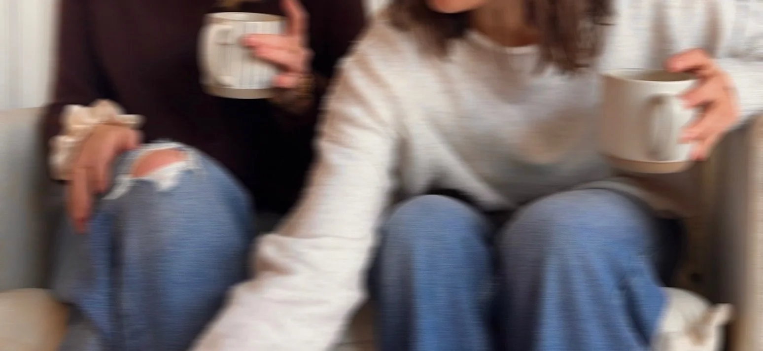 Two people sitting on a couch, each holding a mug, with one person wearing ripped jeans and the other wearing a white sweater.