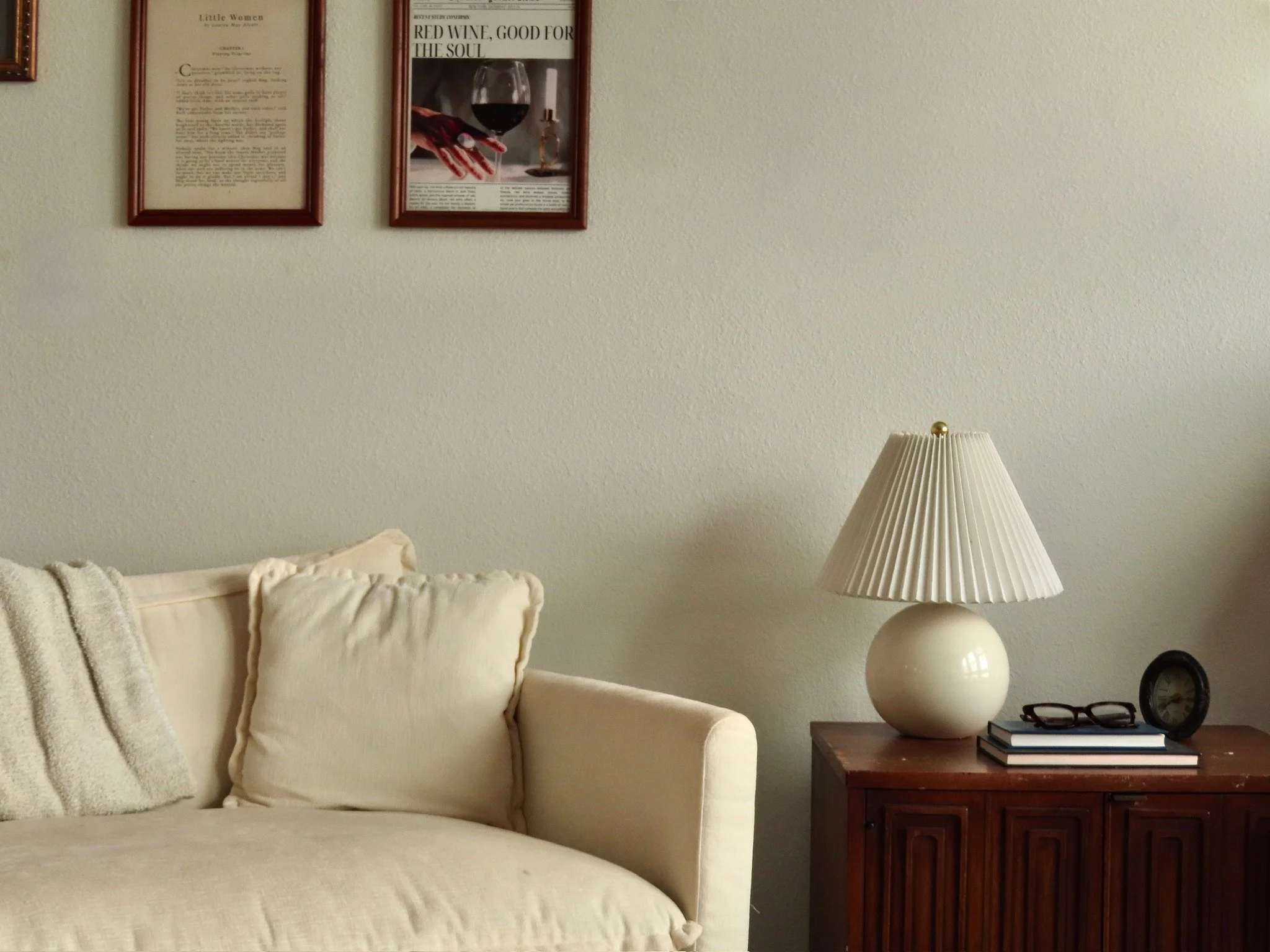 Living room corner with cream sofa, pillows, wooden side table with a large cream lamp, glasses, clock, and books, and framed artwork on a beige wall.