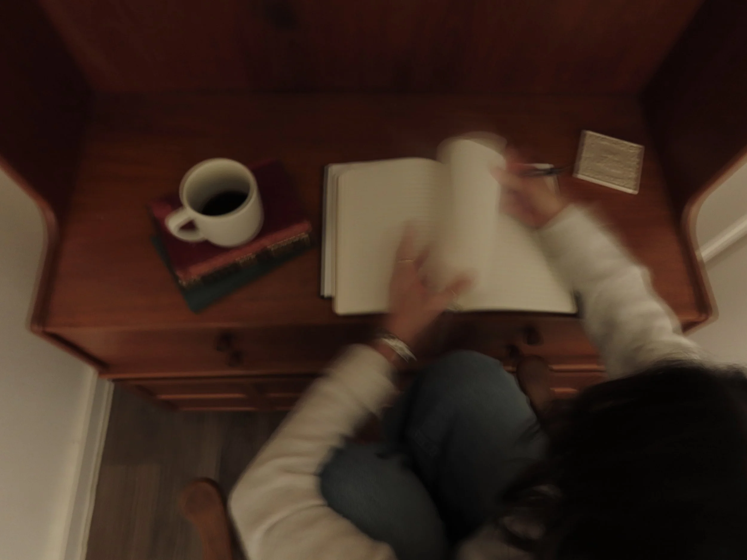 Person sitting at a wooden desk, writing in an open notebook. On the desk are a white coffee mug on top of some books and a small rectangular object. The person is wearing a white long-sleeved shirt and dark pants, and their head is partially visible in the image.