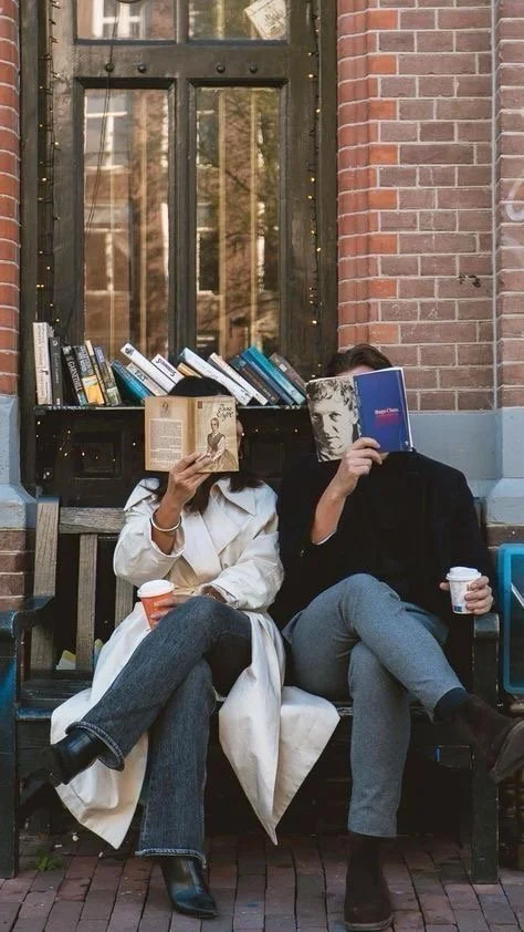 Two people sitting on a bench in front of a brick building with a window and books, each reading a book with their faces hidden behind the books they are holding. They are holding cups and appear to be enjoying themselves.