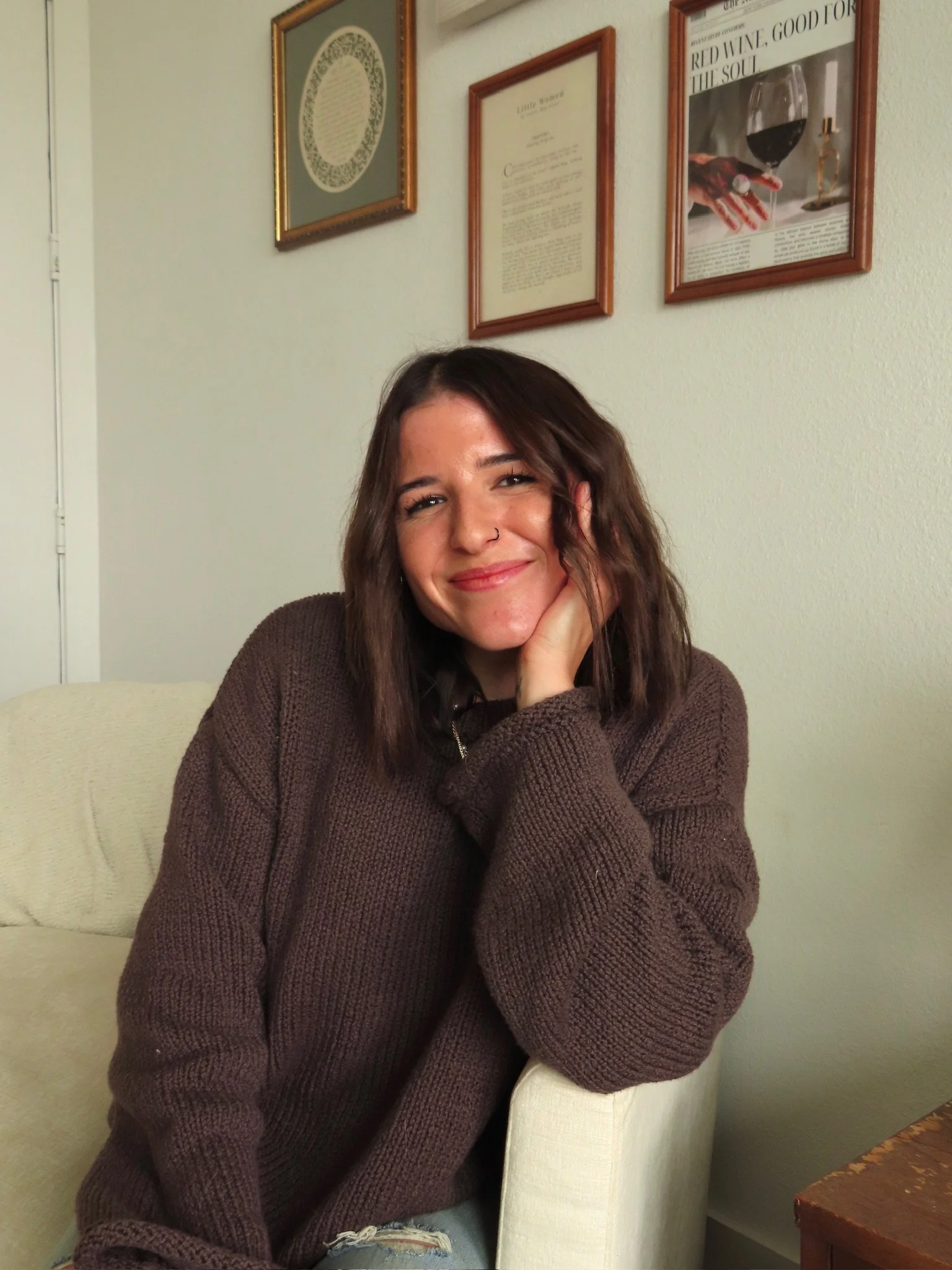 A young woman with shoulder-length brown hair, smiling, sitting on a white armchair, wearing a brown knitted sweater, with framed pictures on the wall behind her.