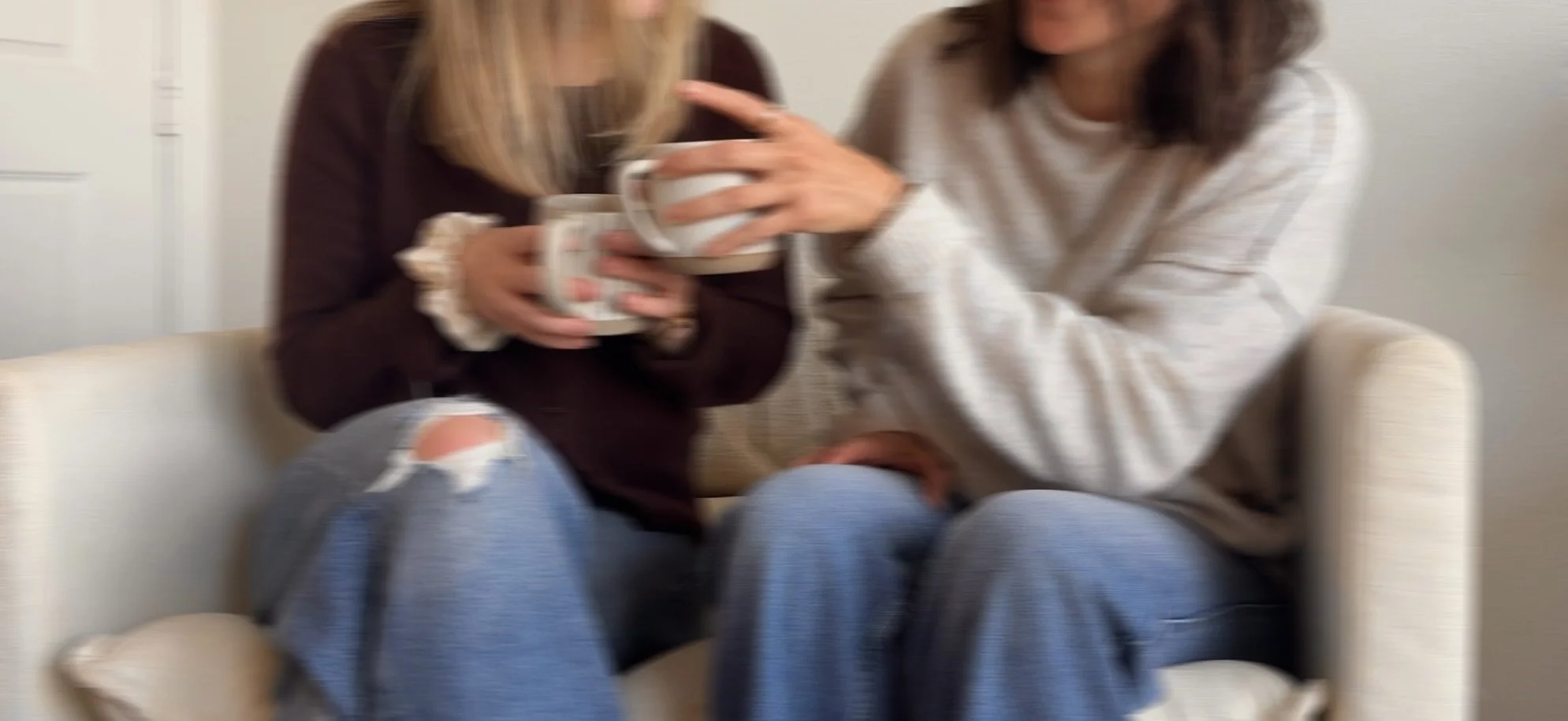 Two women sitting on a cream-colored couch, each holding a white mug, sharing a moment of coffee companionship.