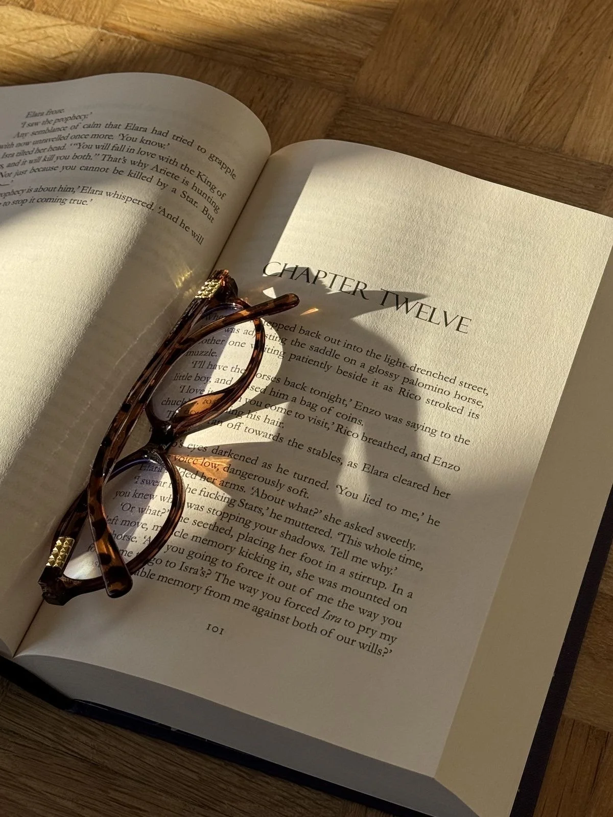 Open book resting on a wooden surface with a pair of tortoiseshell eyeglasses on top, showing Chapter Twelve.
