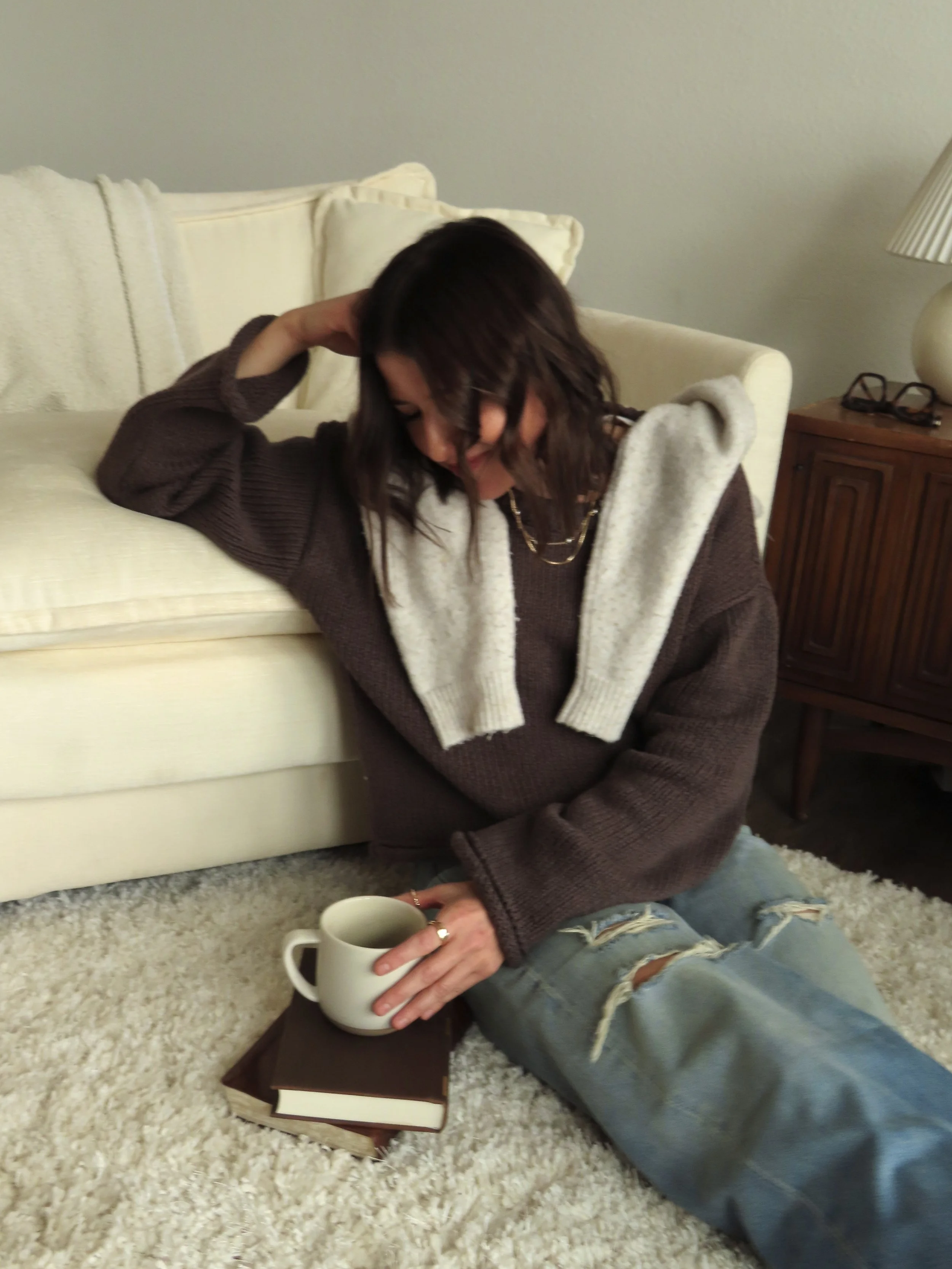 A woman sitting on the carpeted floor with a mug, two books, wearing a brown sweater and ripped jeans, with a sweater draped over her shoulders, in a cozy room.