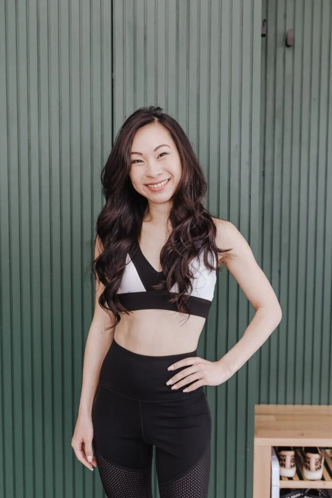 Young woman with long dark hair smiling, standing in front of a green wooden wall, wearing athletic workout clothes.