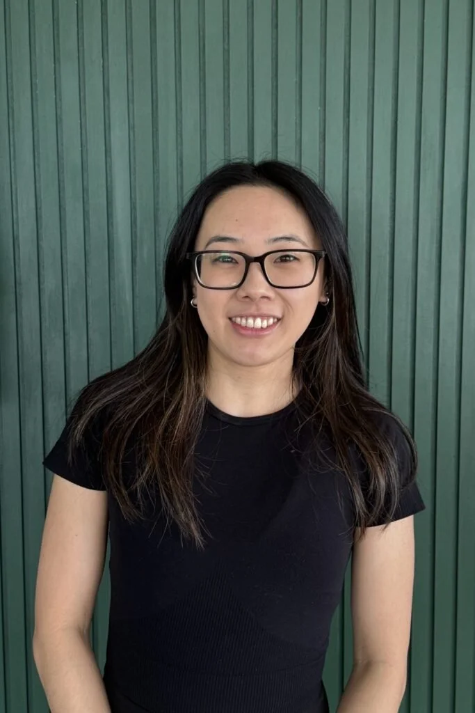 A young woman with long dark hair, glasses, and earrings standing in front of a green vertical panel background, smiling at the camera.