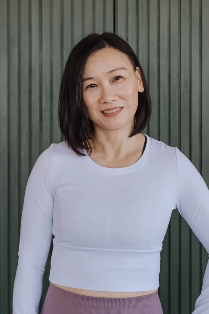 A woman with shoulder-length black hair smiling at the camera, wearing a white long-sleeve crop top and mauve high-waisted pants, standing in front of a green, vertically paneled background.