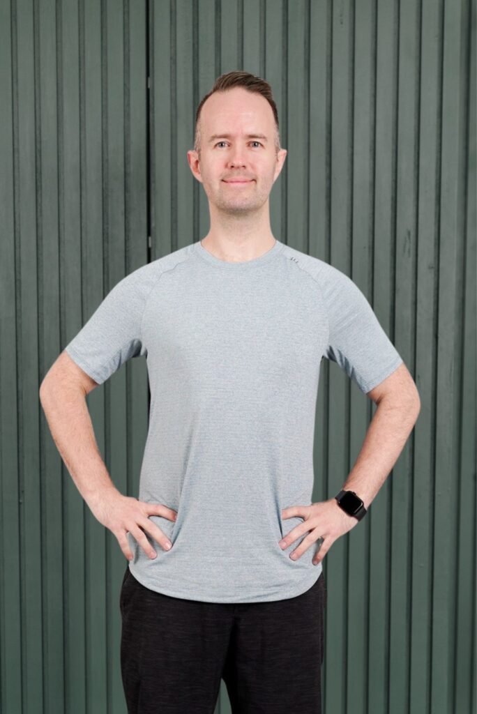 A man with short hair and fair skin posing with hands on his hips in front of a dark green wooden fence, wearing a light gray athletic shirt and black pants, with a smartwatch on his left wrist.