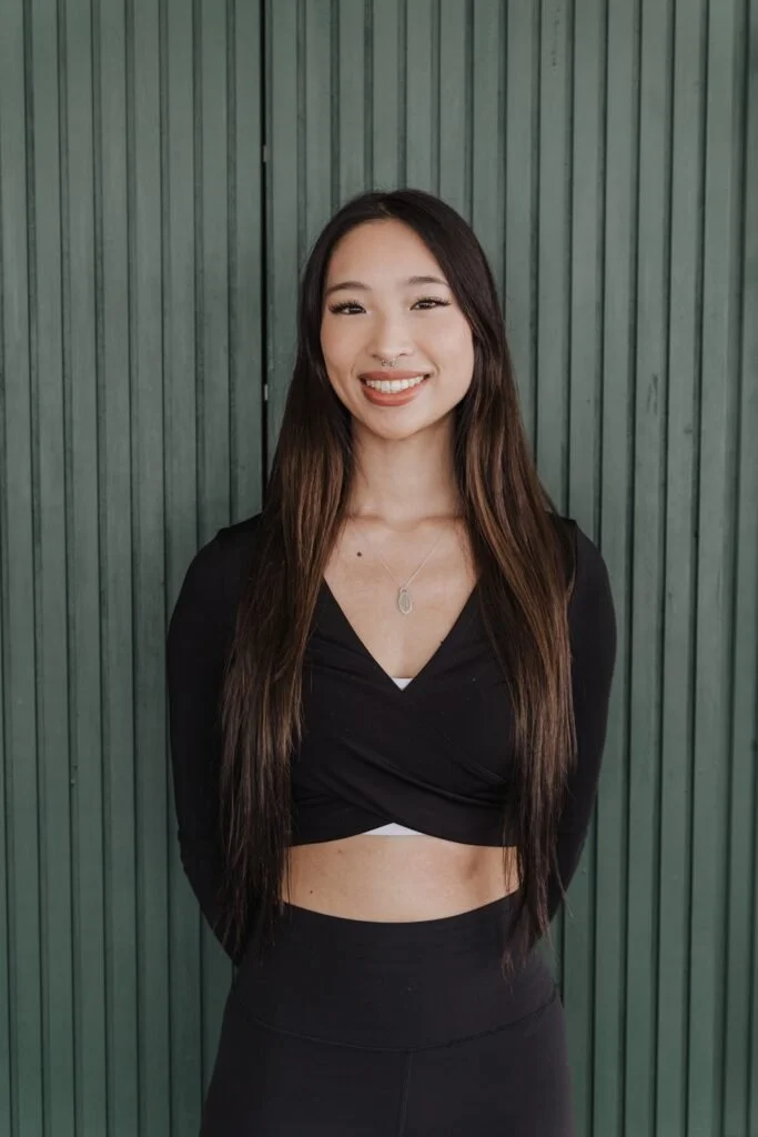 Young woman with long dark hair, wearing a black crop top and black pants, smiling, standing in front of a green wooden panel background.