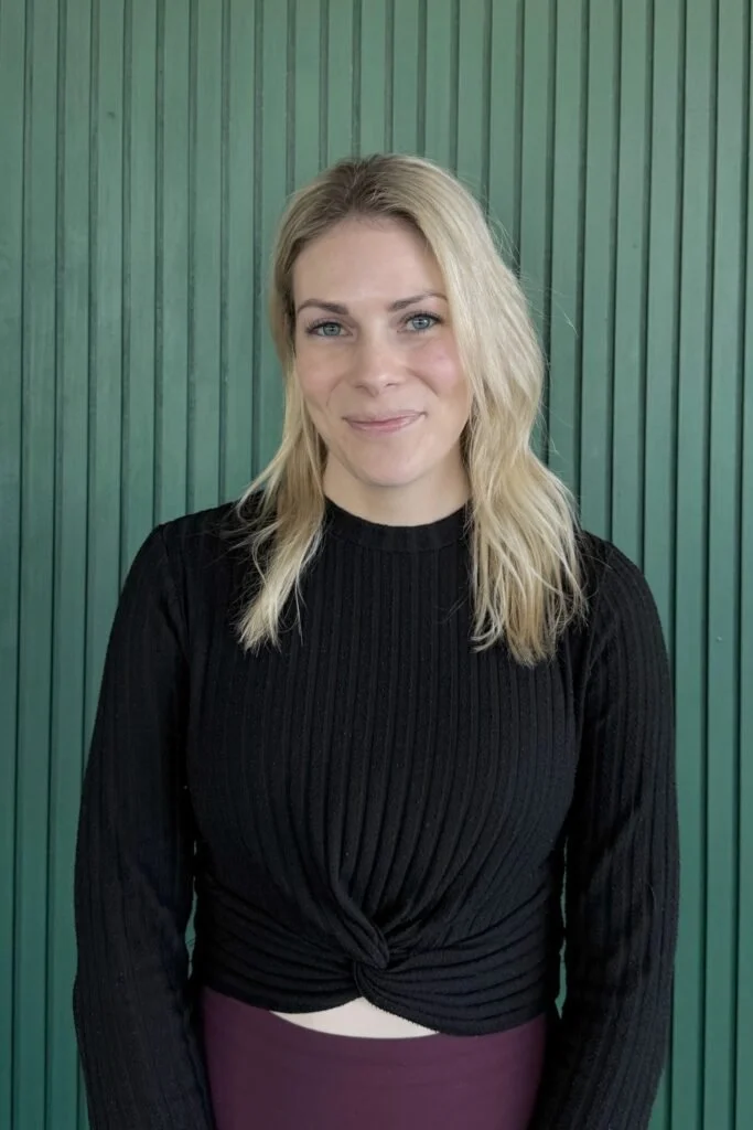 A young woman with blonde hair, blue eyes, and light skin standing in front of a green textured wall, wearing a black long-sleeve ribbed top and maroon skirt.