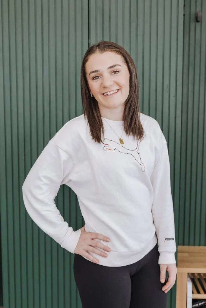 A young woman with shoulder-length brown hair and fair skin, smiling and standing with one hand on her hip. She is wearing a white sweatshirt with a graphic design of a map and a gold necklace. The background is a green vertical wood panel wall.