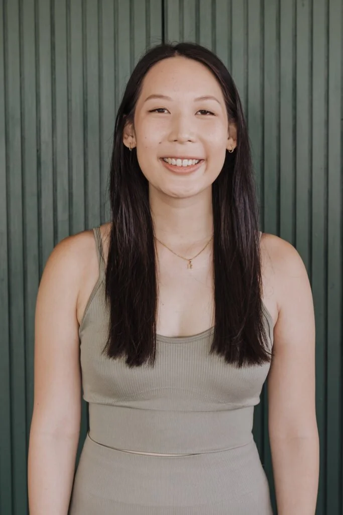 A young woman with long dark hair, smiling and wearing a beige sleeveless top, standing in front of a green wooden wall.